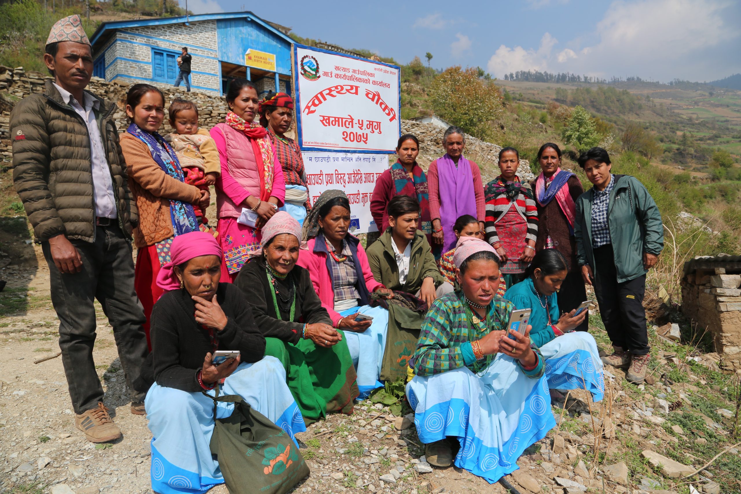 Women seated outside a rural health post in Mugu using mobile phones